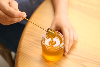 Photo of Woman holding honey dipper over jar at wooden table, closeup