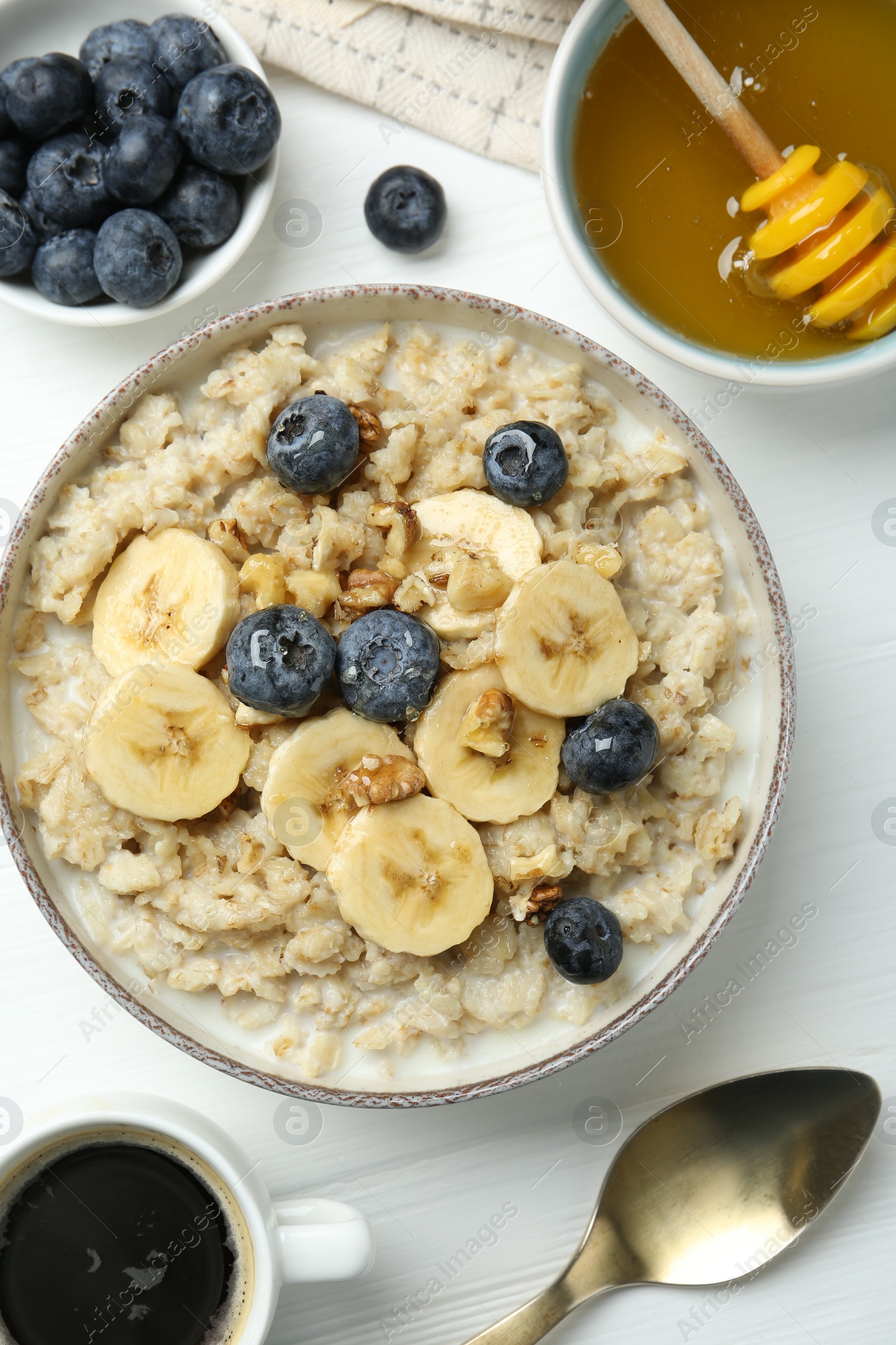 Photo of Tasty oatmeal with banana, blueberries, walnuts and honey served in bowl on white wooden table, flat lay