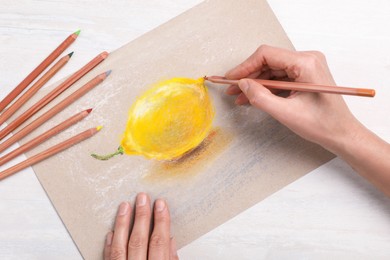 Photo of Woman drawing lemon on paper with pastel pencil at white wooden table, top view