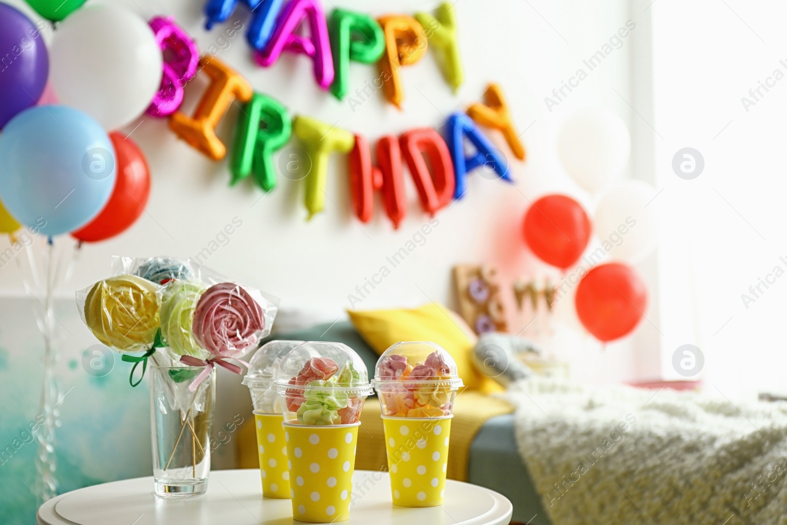 Photo of Table with treats and phrase HAPPY BIRTHDAY made of colorful balloon letters in modern bedroom. Space for text