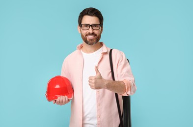 Photo of Architect with drawing tube and hard hat showing thumbs up on light blue background