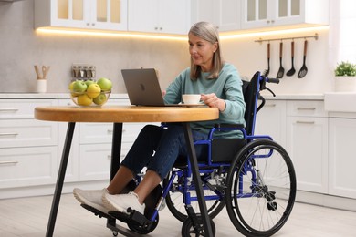 Photo of Woman in wheelchair with cup of drink using laptop at table in home office