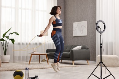 Happy sports blogger jumping with rope while streaming online fitness lesson with smartphone at home