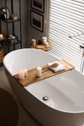 Wooden tray with burning candles, towel and brush on bathtub in bathroom