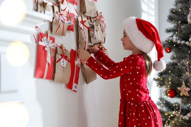 Photo of Cute little girl in Santa hat taking gift from Christmas advent calendar at home