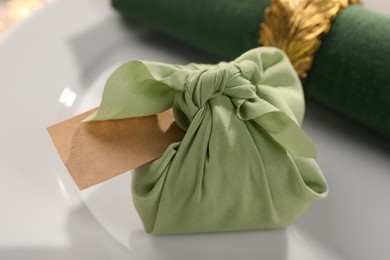 Photo of Furoshiki technique. Gift packed in green fabric, blank card and napkin on plate, closeup