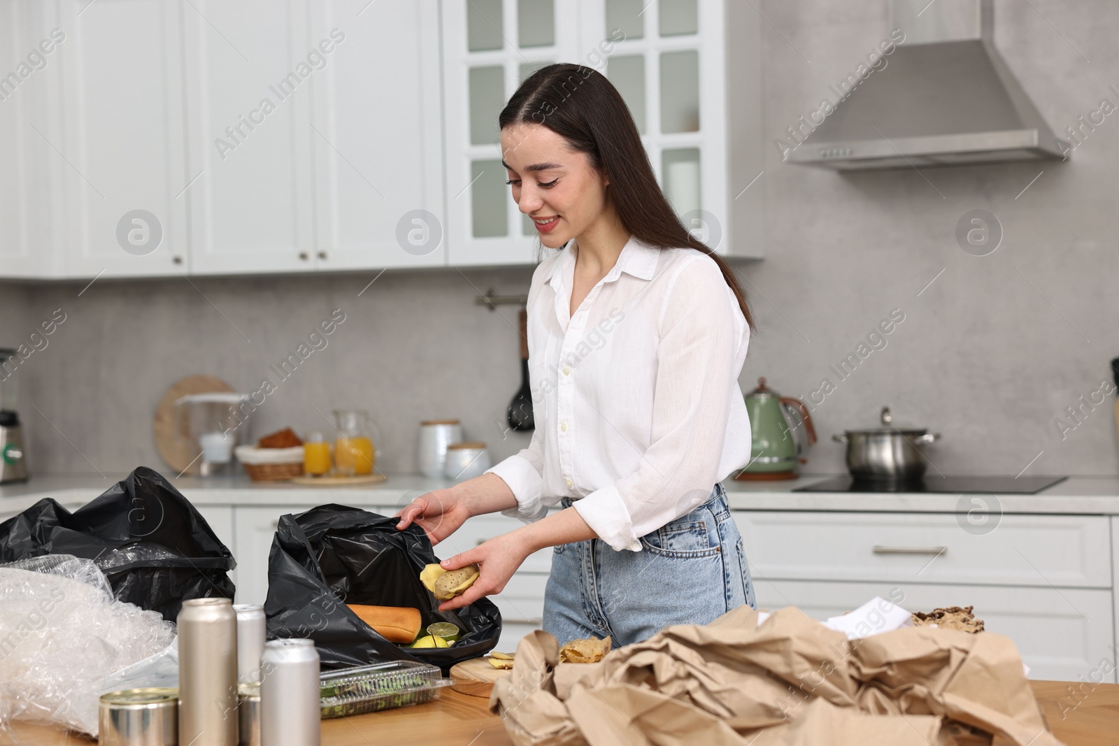 Photo of Garbage sorting. Woman putting food waste into plastic bag at table in kitchen