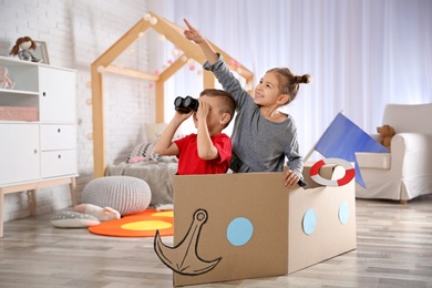 Photo of Cute little kids playing with binoculars and cardboard boat in bedroom