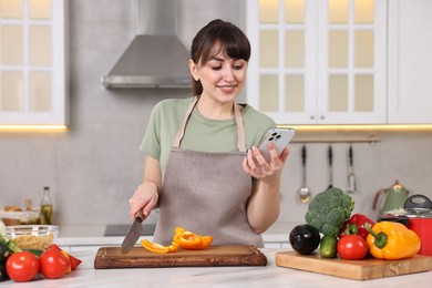Photo of Happy young housewife using smartphone while cutting bell pepper at white marble table in kitchen