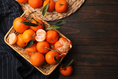Photo of Fresh ripe tangerines with green leaves in wicker basket on wooden table, flat lay. Space for text