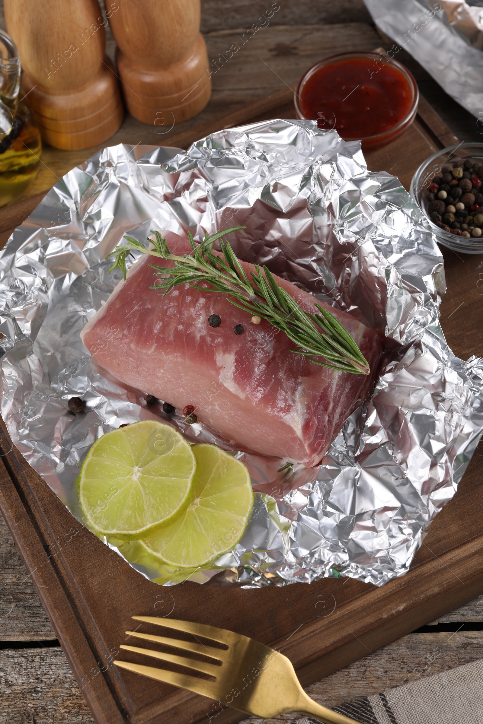 Photo of Aluminum foil with raw meat, rosemary, lime and spices on wooden table, above view