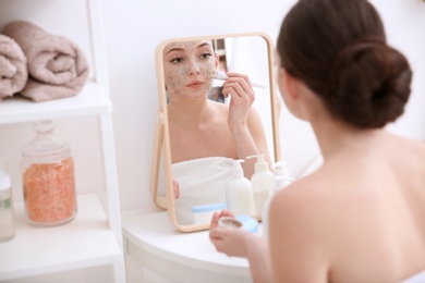 Photo of Woman applying scrub onto face in front of mirror at home