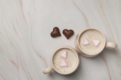 Cups of aromatic coffee with heart shaped marshmallows and chocolate candies on white marble table, flat lay. Space for text