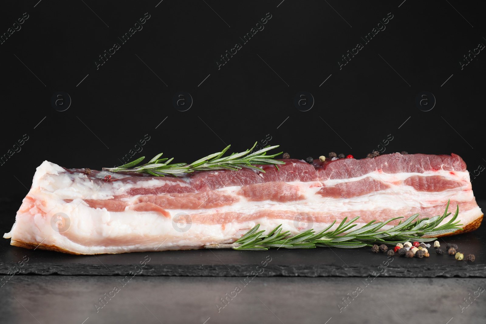 Photo of Piece of raw pork belly, rosemary and peppercorns on grey table
