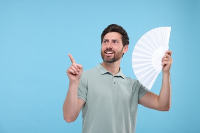 Happy man holding hand fan on light blue background. Space for text