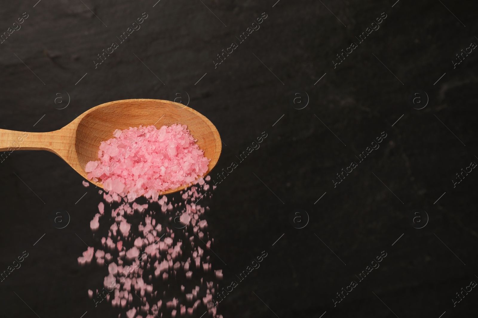 Photo of Pouring pink sea salt from wooden spoon against black background, closeup. Space for text