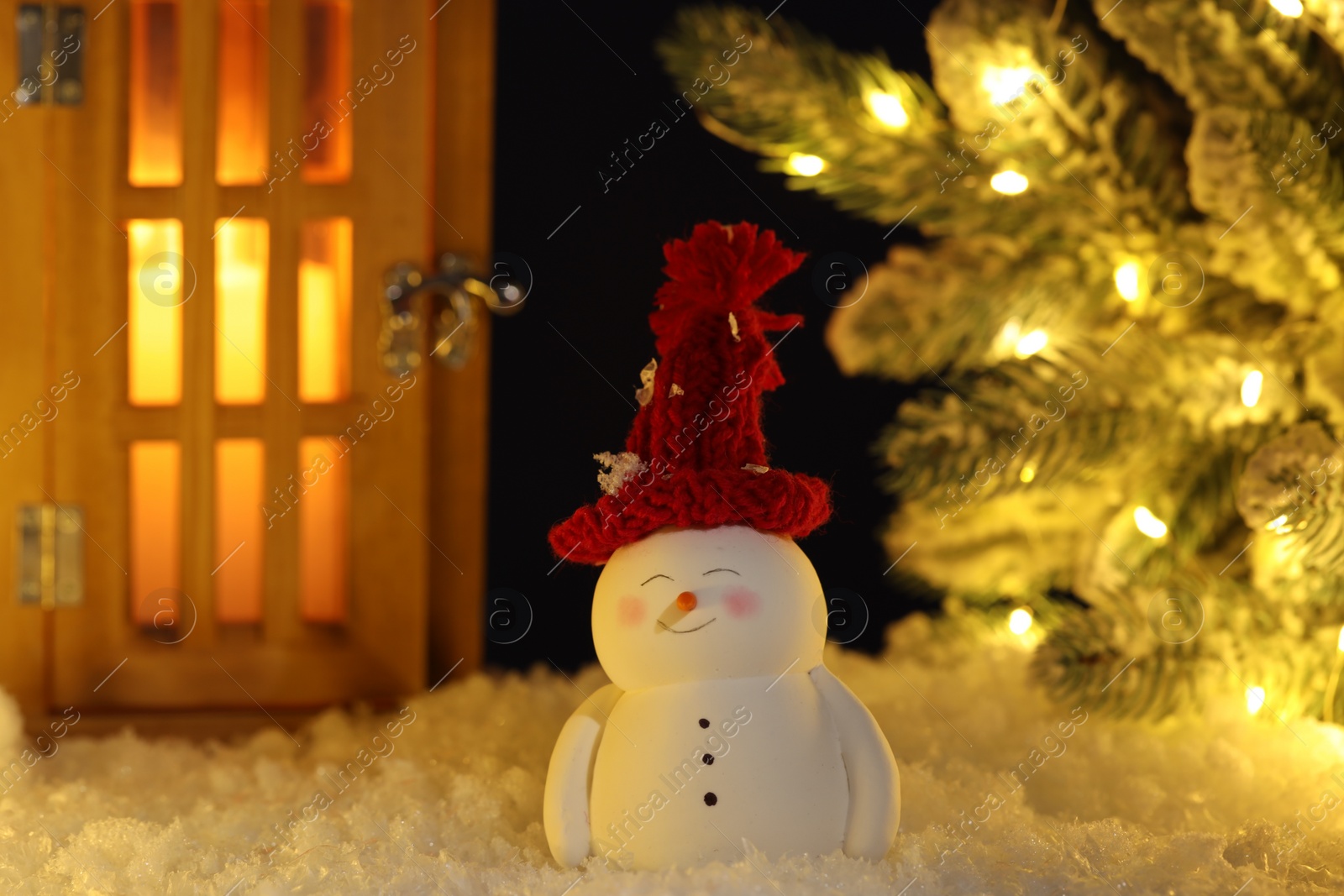 Photo of Cute decorative snowman, lantern and Christmas tree on artificial snow against dark background