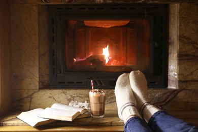 Woman with glass of hot cocoa and books near fireplace at home, closeup