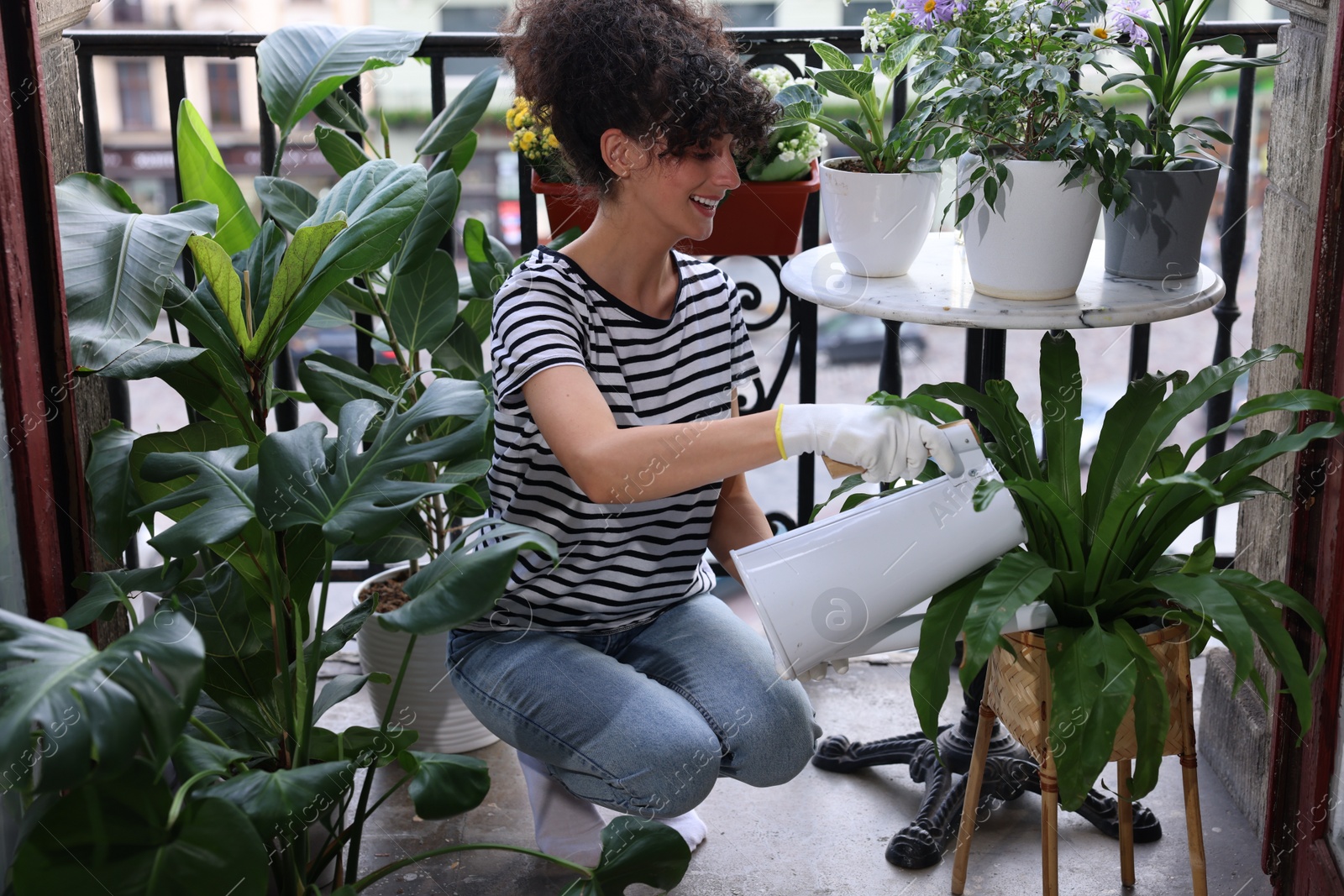Photo of Happy young woman watering green potted houseplants on balcony