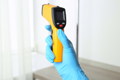 Photo of Doctor in gloves holding non contact infrared thermometer indoors, closeup