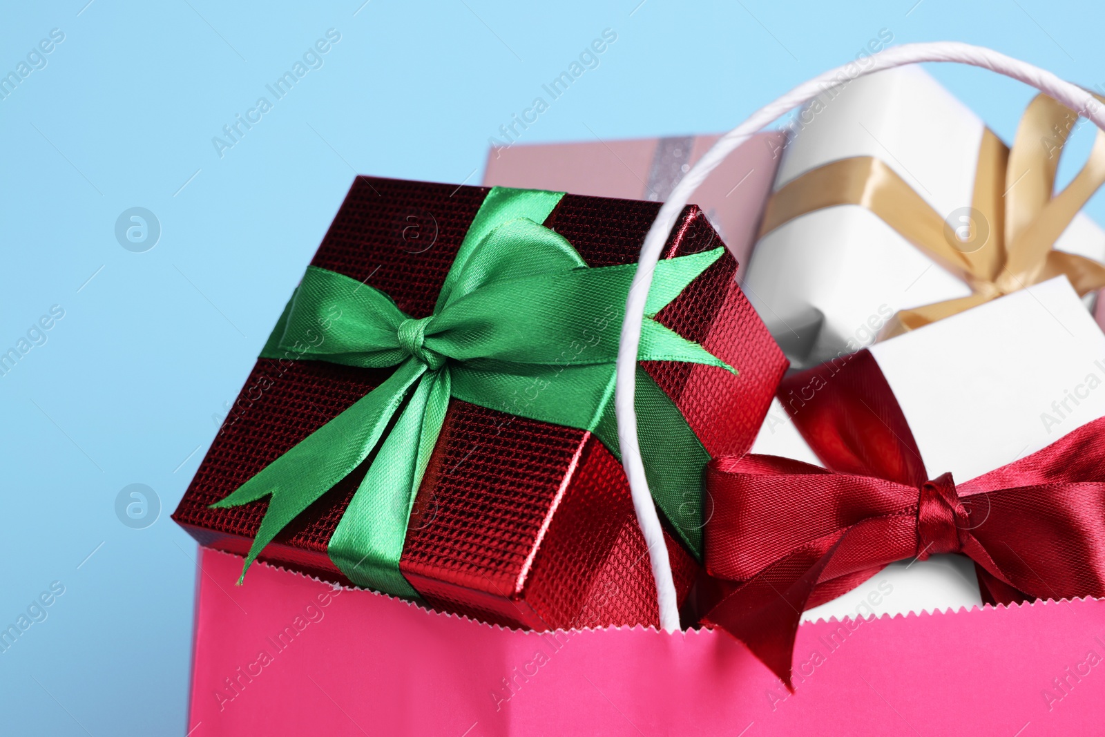 Photo of Pink paper shopping bag full of gift boxes on light blue background, closeup