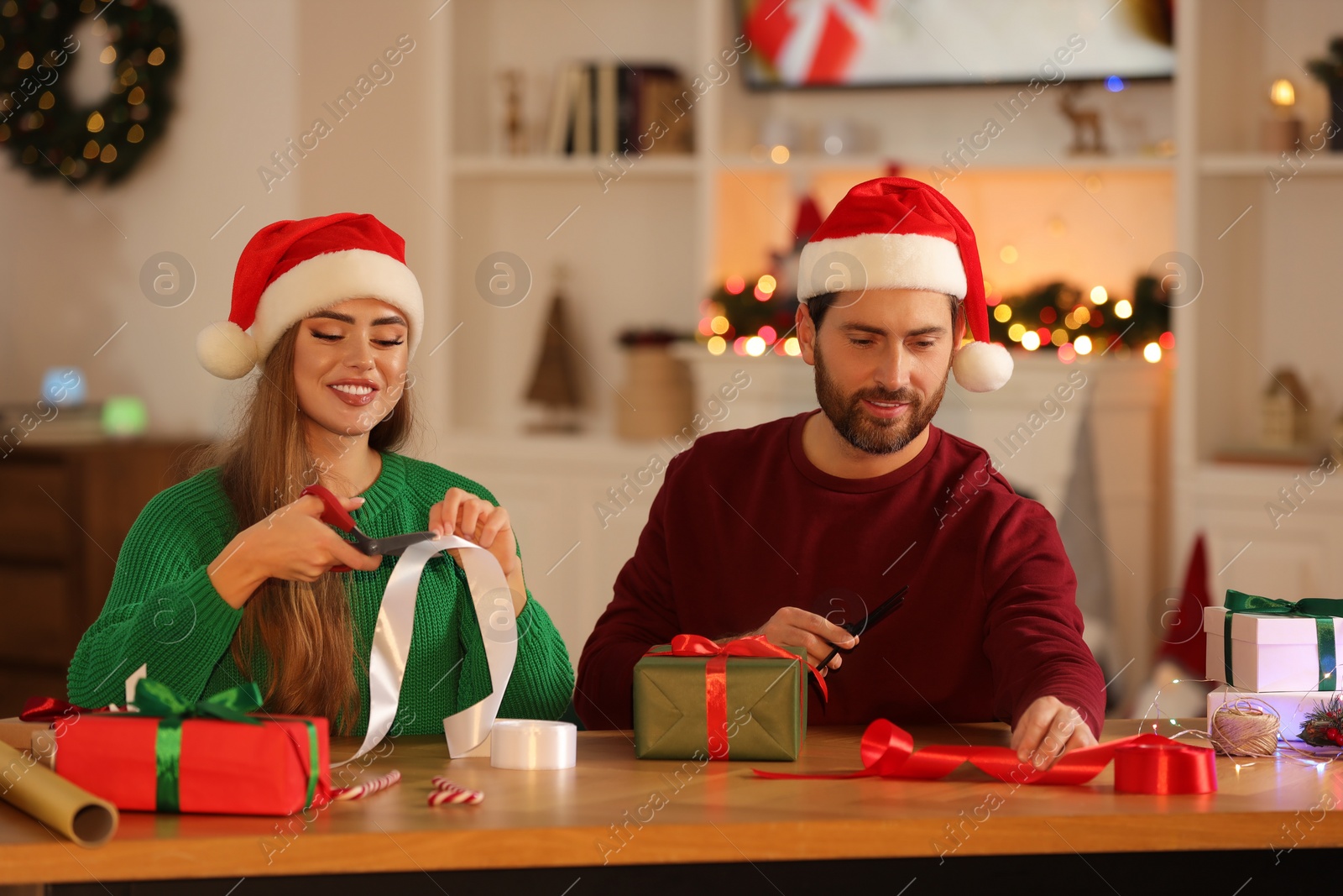 Photo of Happy couple in Santa hats decorating Christmas gifts at table in room