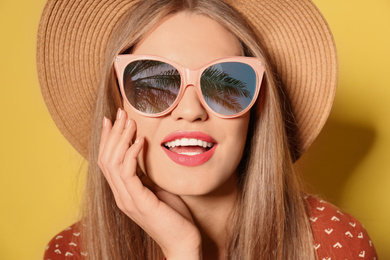 Young woman wearing stylish sunglasses with reflection of palm trees and hat on yellow background 