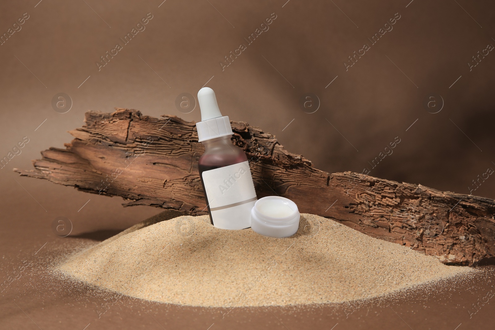 Photo of Cosmetic products and tree bark on sand against brown background