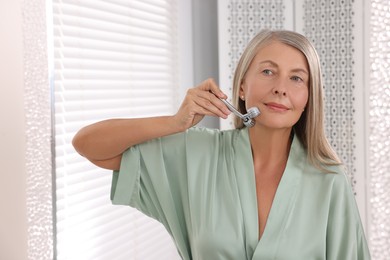 Photo of Woman massaging her face with metal roller near mirror in bathroom