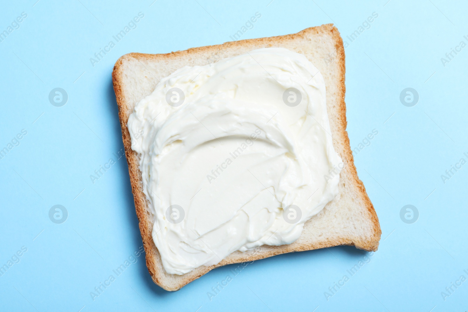 Photo of Slice of bread with tasty cream cheese on light blue background, top view