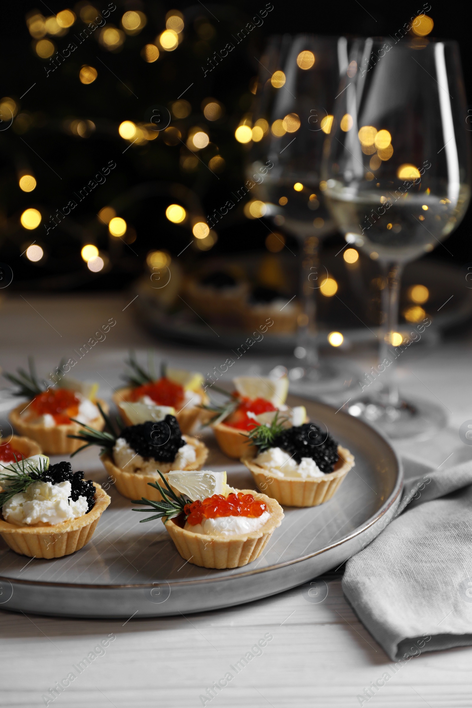 Photo of Delicious tartlets with red and black caviar served on white wooden table against blurred festive lights, closeup. Space for text