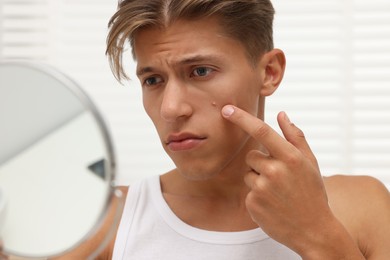 Photo of Upset young man looking at mirror and touching pimple on his face indoors. Acne problem