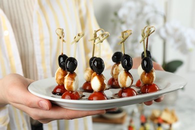 Photo of Woman holding plate of tasty canapes with black olives, mozzarella and cherry tomatoes indoors, closeup