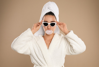 Photo of Young woman in bathrobe and sunglasses blowing chewing gum on brown background