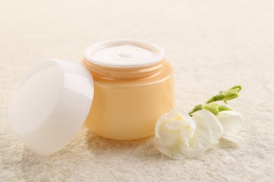 Photo of Body care. Moisturizing cream in open jar and flower on light textured table, closeup