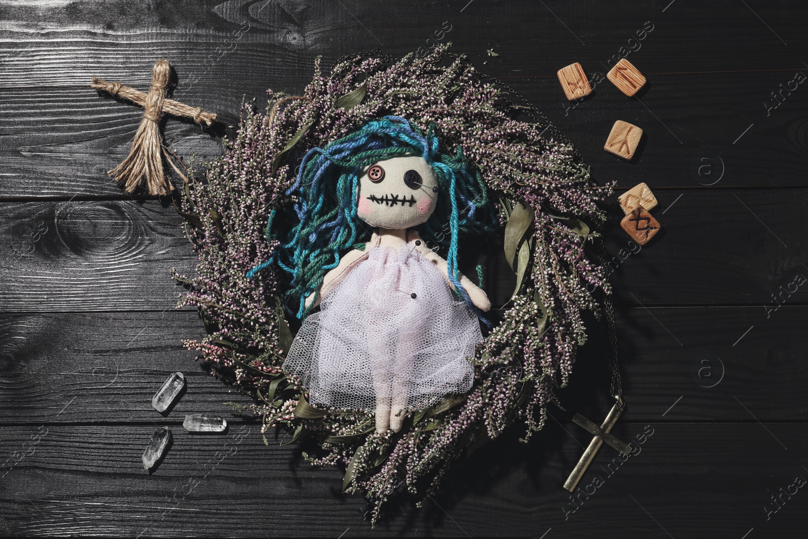 Photo of Female voodoo doll with pins surrounded by ceremonial items on black wooden background, flat lay