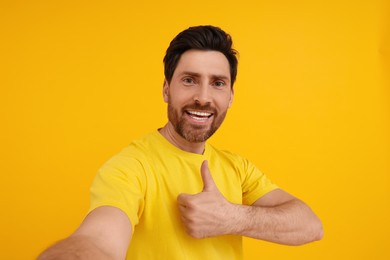 Photo of Smiling man taking selfie and showing thumbs up on yellow background