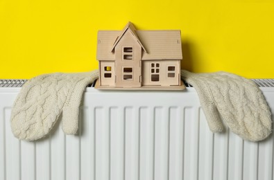 Photo of Wooden house model and knitted mittens on heating radiator near yellow wall. Energy efficiency concept
