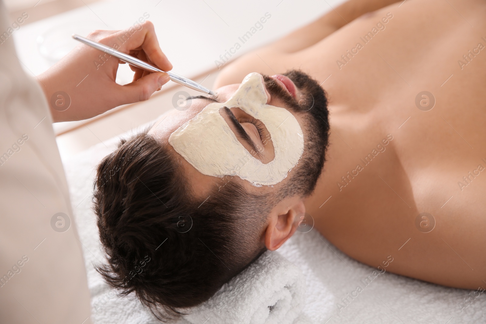 Photo of Cosmetologist applying mask on man's face in spa salon