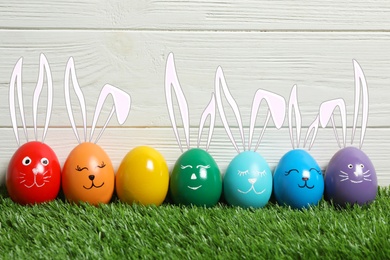 Colorful eggs as Easter bunnies on green grass against wooden background