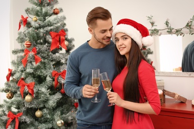 Photo of Young beautiful couple with glasses of champagne near Christmas tree at home