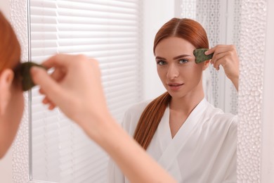 Photo of Young woman massaging her face with jade gua sha tool near mirror in bathroom