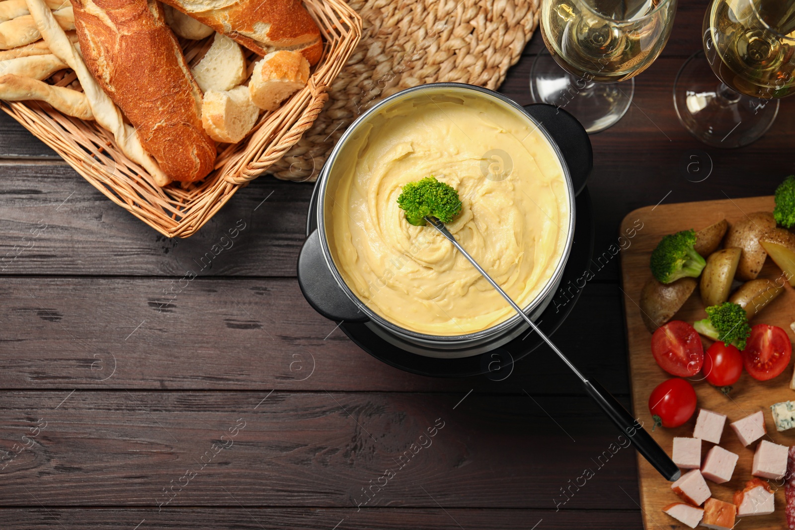 Photo of Fork with piece of broccoli, melted cheese in fondue pot, wine and snacks on wooden table, flat lay. Space for text