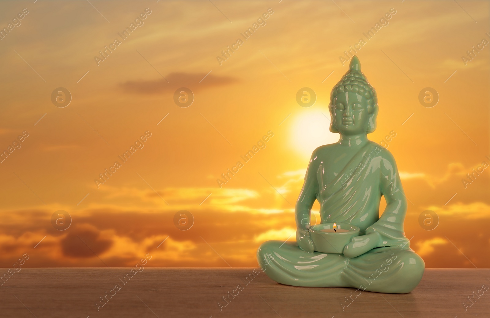 Image of Beautiful ceramic Buddha sculpture with burning candle on wooden surface at sunset. Space for text