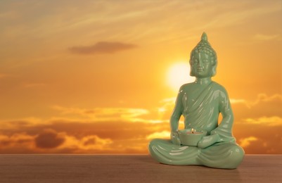 Image of Beautiful ceramic Buddha sculpture with burning candle on wooden surface at sunset. Space for text