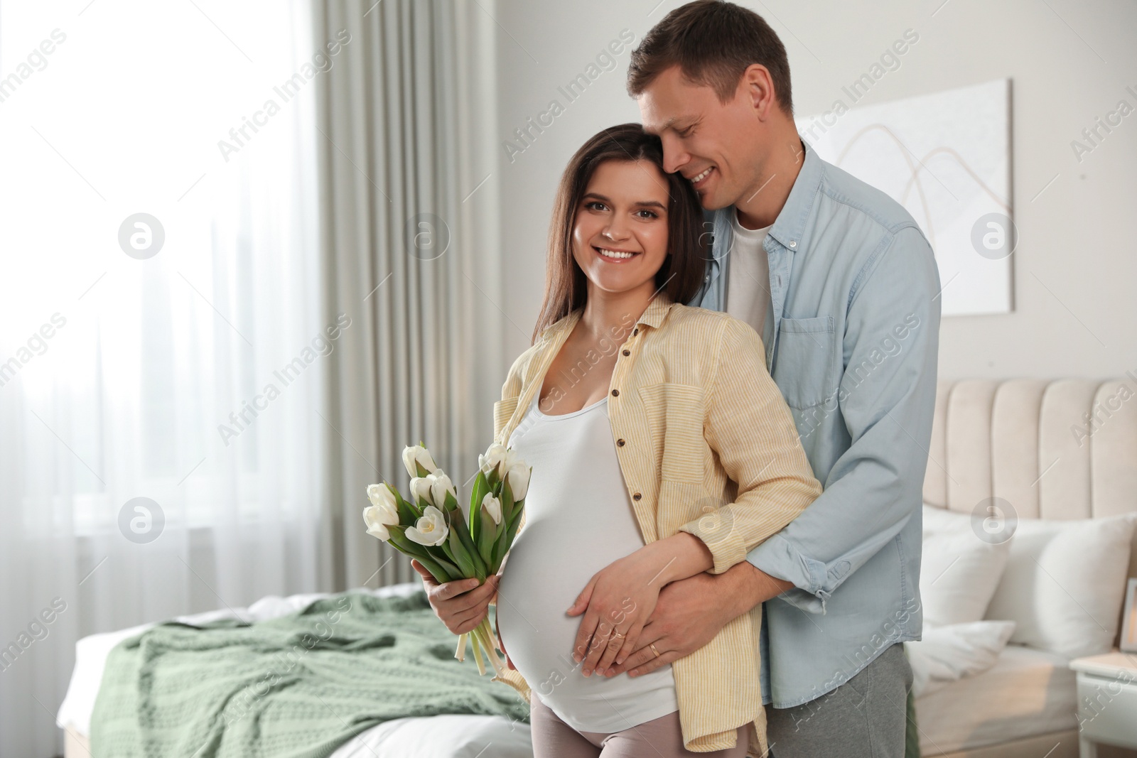 Photo of Young pregnant woman with flowers and her husband in bedroom