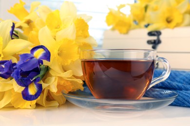 Cup of aromatic tea, beautiful yellow daffodil and iris flowers on white table