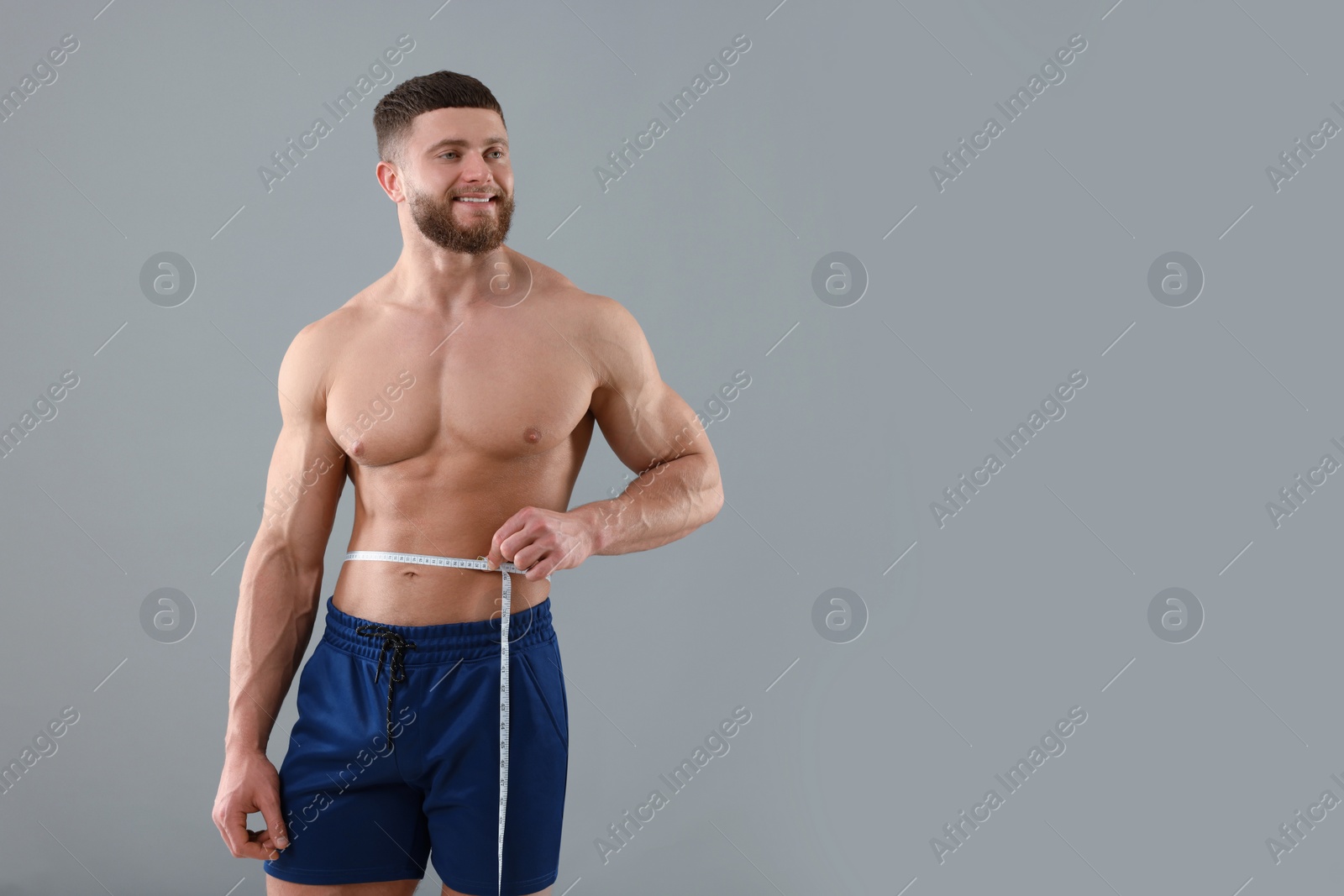 Photo of Portrait of happy athletic man measuring waist with tape on grey background, space for text. Weight loss concept