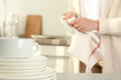 Photo of Woman wiping plate with towel in kitchen, focus on stack of clean dishes