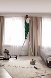 Photo of Professional janitor on ladder near window with curtains in bedroom. Cleaning service